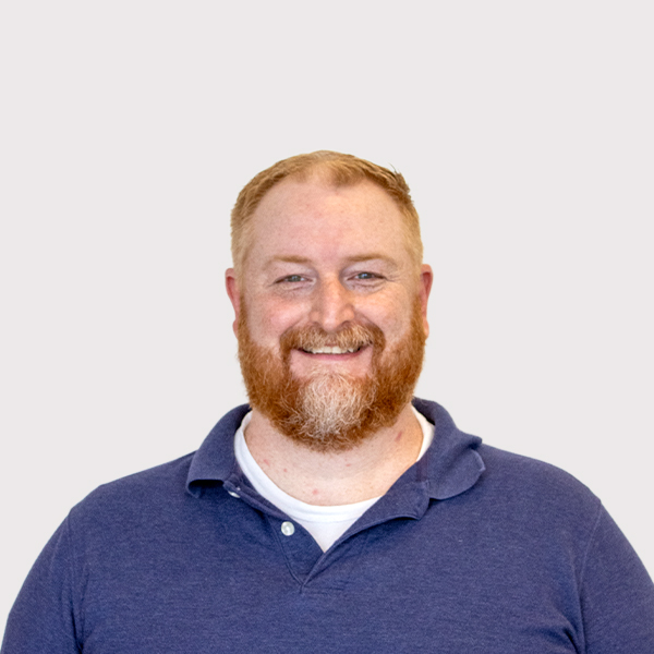 Professional headshot of a smiling man with a beard, wearing a blue polo shirt against a light gray background.